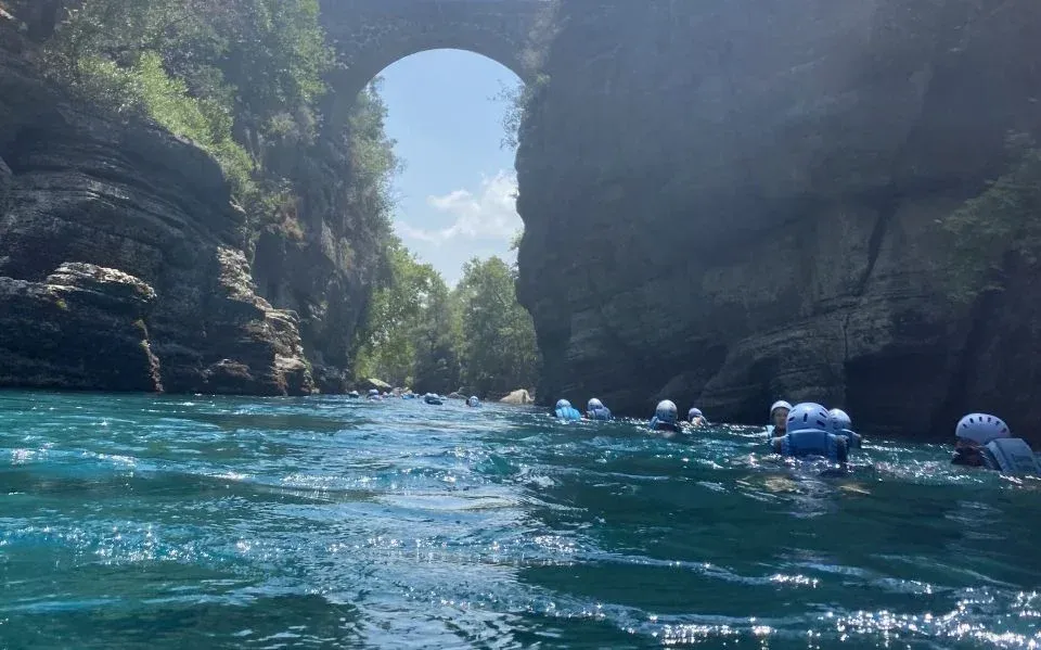 The Roman Oluk Bridge arching high over the turquoise Köprüçay river, with helmeted rafters floating downstream